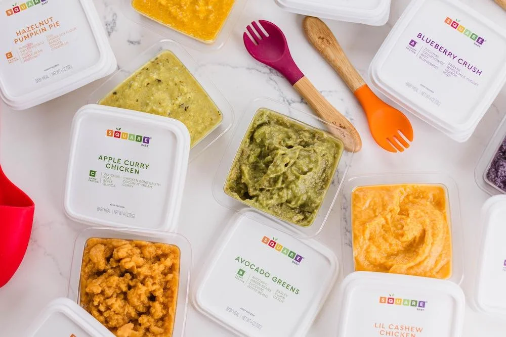 Assorted containers of baby food including avocado greens, apple curry chicken, blueberry crush, hazelnut pumpkin pie, and others on a white surface with colorful utensils.