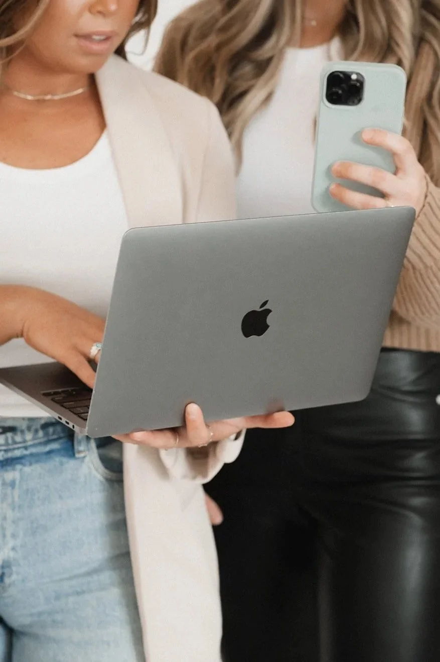 Two women are looking at a MacBook laptop and taking a selfie with a smartphone. One woman is wearing a white top and a beige cardigan, and the other is dressed in black leather pants and a beige sweater.