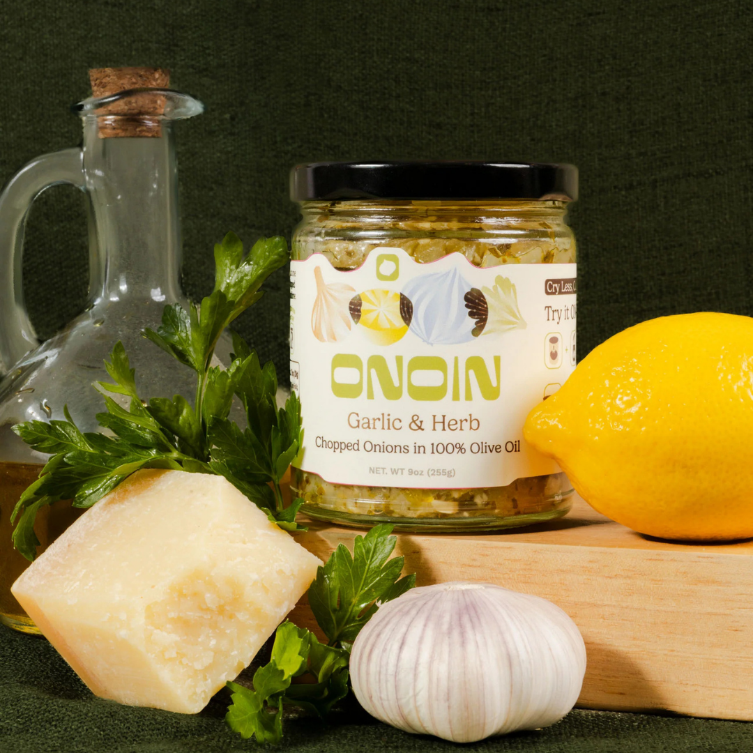 A jar labeled 'ONION Garlic & Herb' containing chopped onions in olive oil, a whole lemon, a bulb of garlic, fresh parsley, and a glass bottle of olive oil on a wooden surface against a dark background.