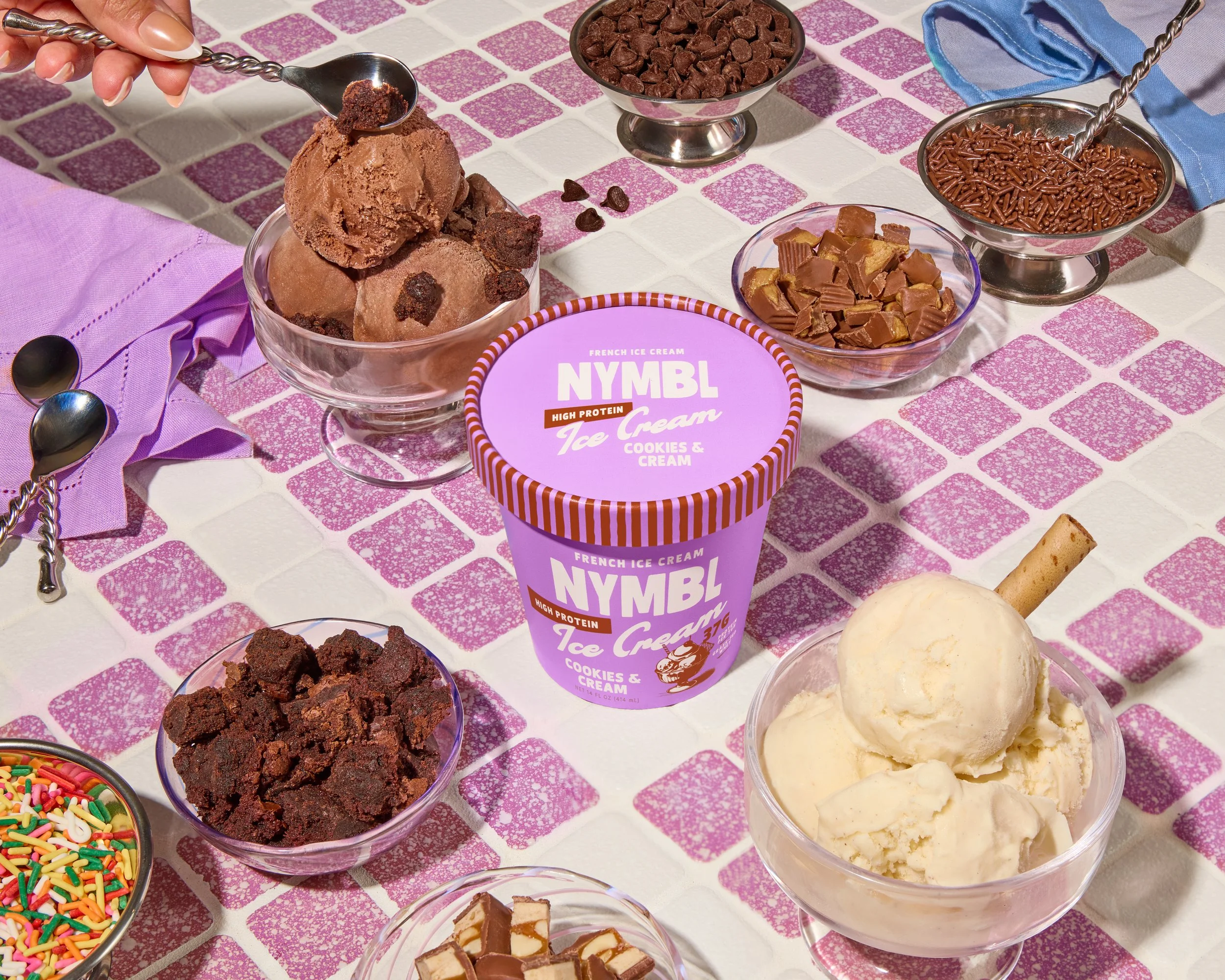 Various bowls of ice cream flavors including chocolate chip, vanilla, and chocolate, along with bowls of toppings such as sprinkles, chocolate chunks, crushed cookies, and chocolate sprinkles, arranged on a pink and white tiled table.