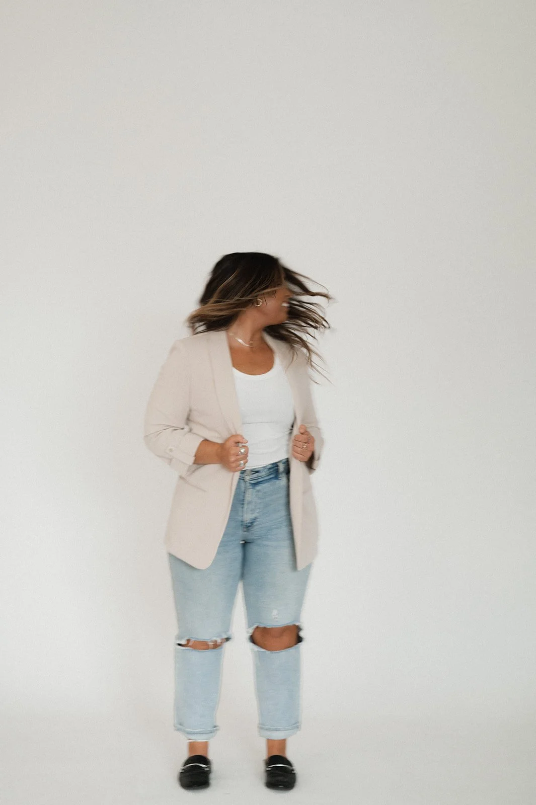 Woman with brown hair wearing a beige blazer, white t-shirt, ripped jeans, and black shoes, posing against a plain white background.