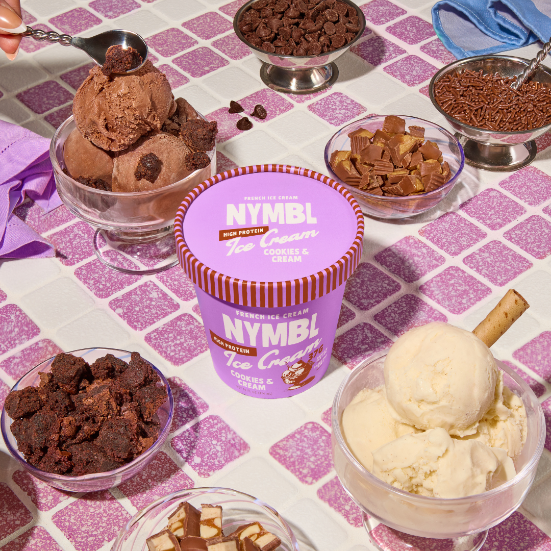 An assortment of ice cream flavors in bowls surrounding a purple container labeled 'NYMB Ice Cream Cookies & Cream,' on a pink and white tiled table. The flavors include chocolate, caramel, brownie, and vanilla with a cookie stick.