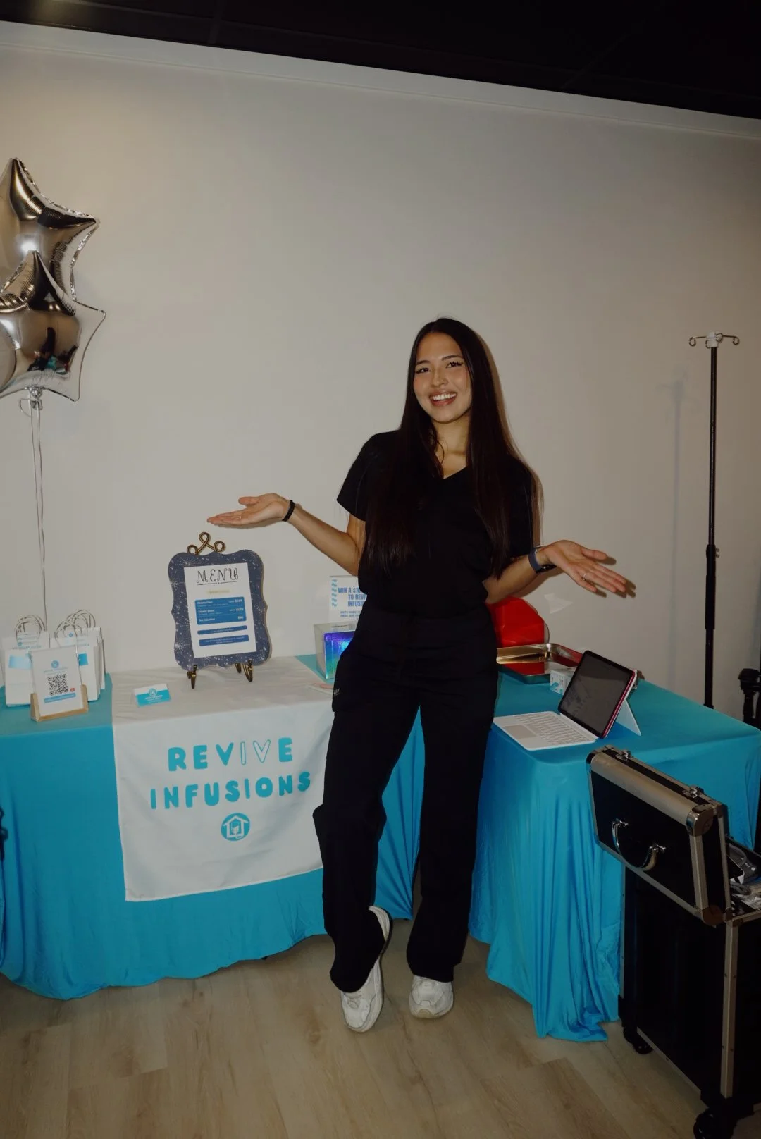Smiling woman standing at a Revive Infusions event booth with a menu sign, blue tablecloths, promotional materials, and a laptop.