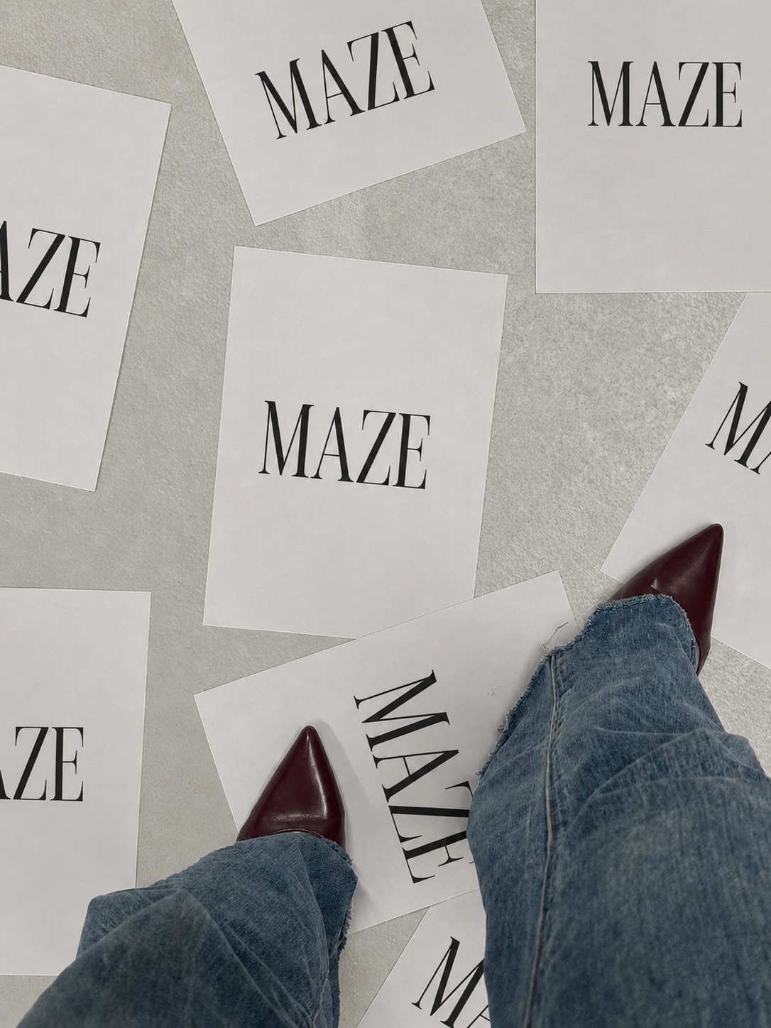 View of person's legs wearing jeans and maroon boots standing on scattered papers with the word "MAZE" printed on them, on a light-colored floor.