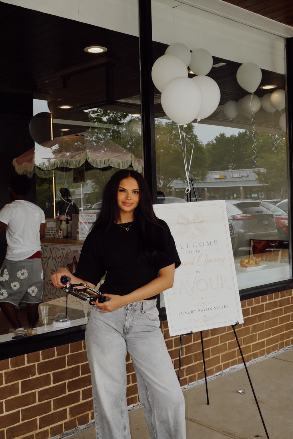 Woman standing outside a salon grand opening with white balloons and a welcome sign
