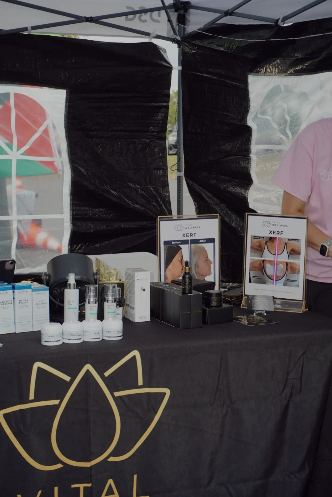 Vital Wellness booth displaying skincare products, XERF treatment brochures, and promotional before-and-after photos at an outdoor market