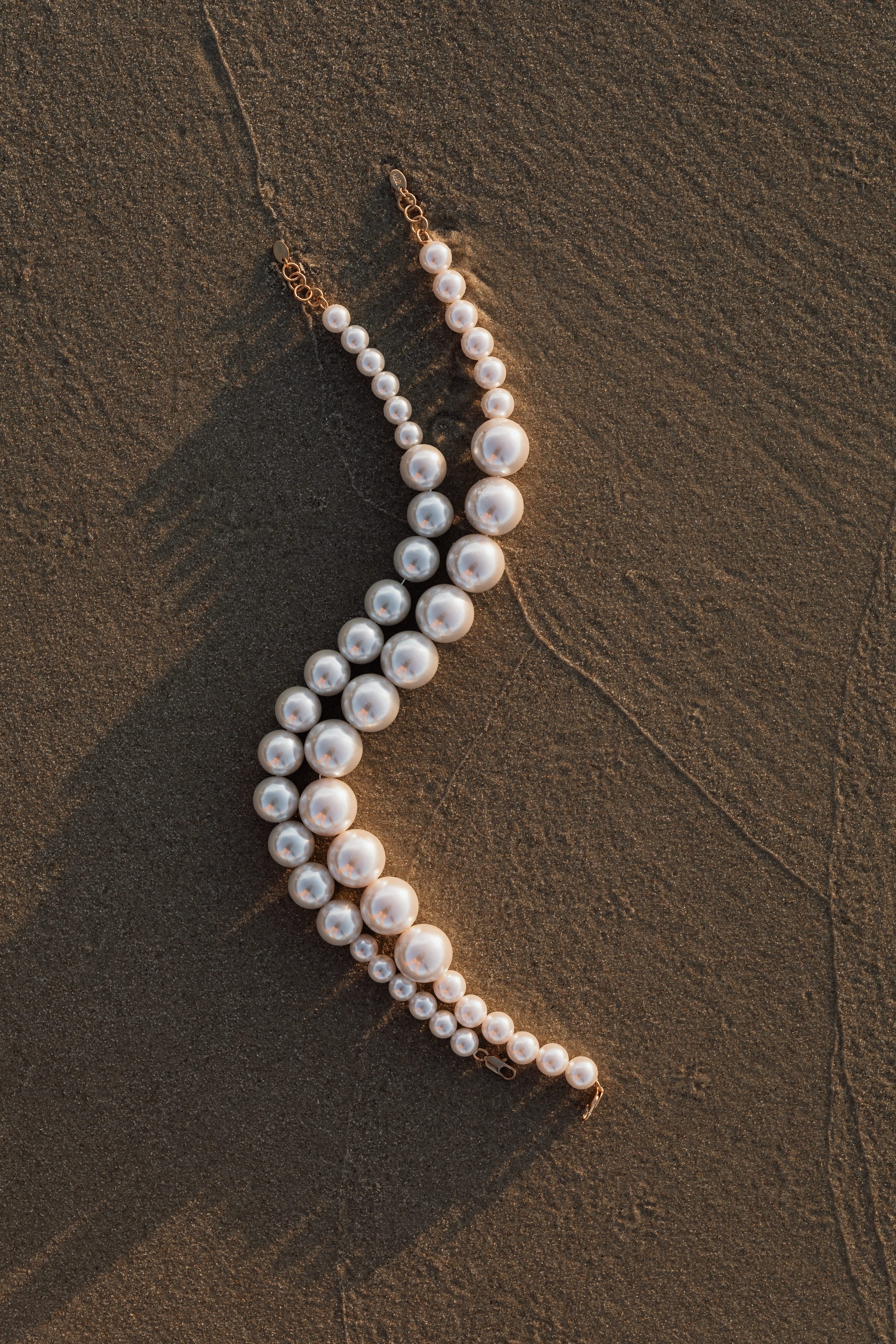 A pearl necklace with a gold chain and clasp laying on dark textured sand.