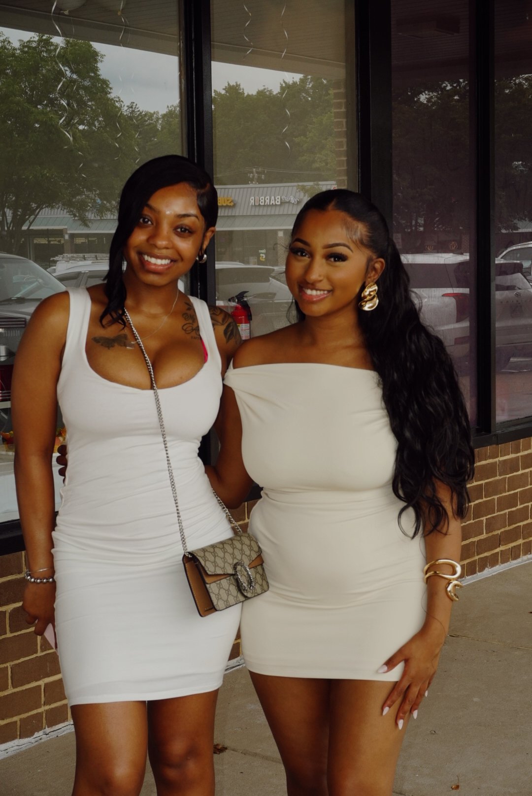 Two women smiling in white dresses outside a storefront