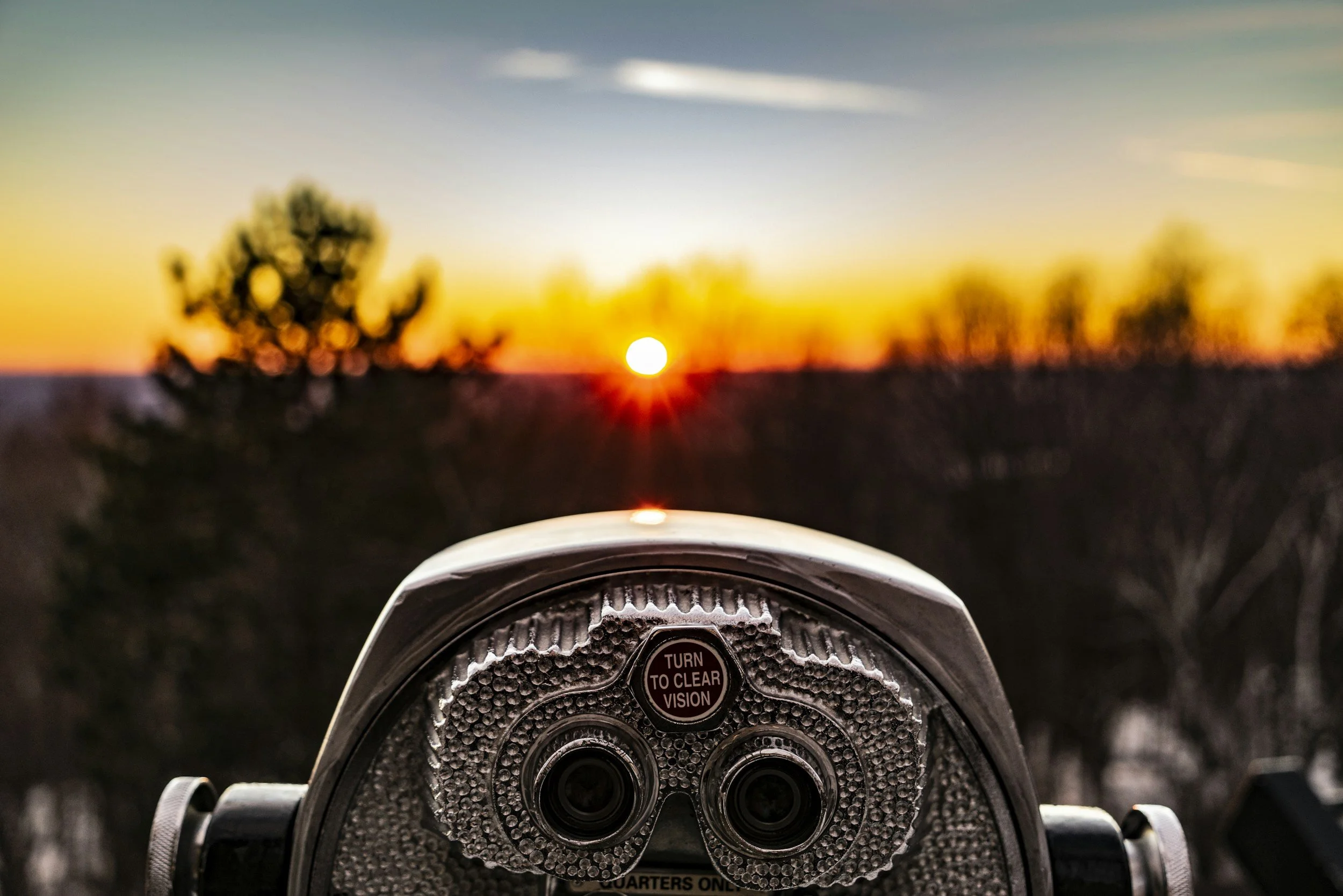 Close-up of a coin-operated viewing binoculars pointed at a sunset scene with blurred trees and sky.