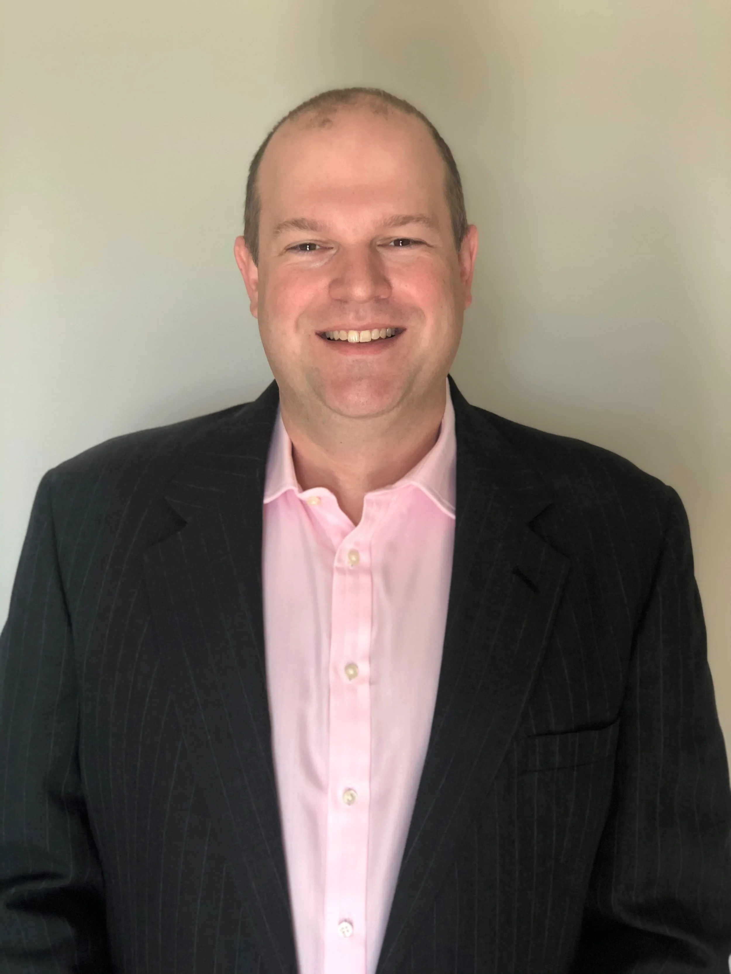 Headshot of a smiling man with short hair, wearing a dark suit jacket and a light pink dress shirt, standing against a plain light-colored wall.