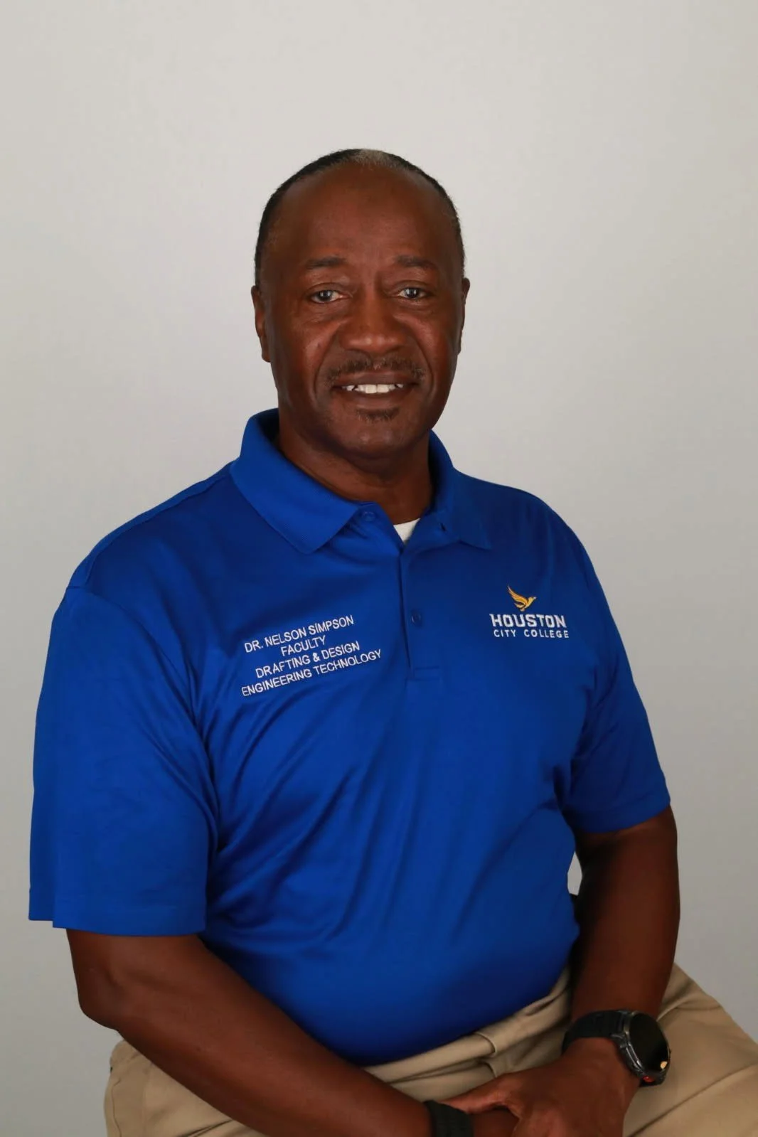A middle-aged African American man wearing a blue polo shirt with university logos and text, sitting against a plain white wall.
