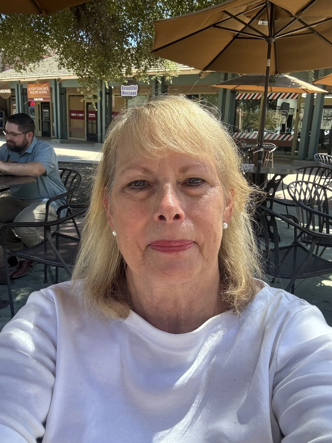 Close-up of a woman with blonde hair, wearing a white top and earrings, sitting outdoors at a cafe with umbrellas, surrounded by chairs and tables.