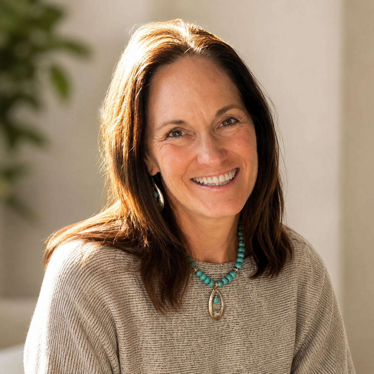 A smiling woman with shoulder-length brown hair, wearing a beige sweater and turquoise jewelry, sitting indoors with sunlight shining on her.
