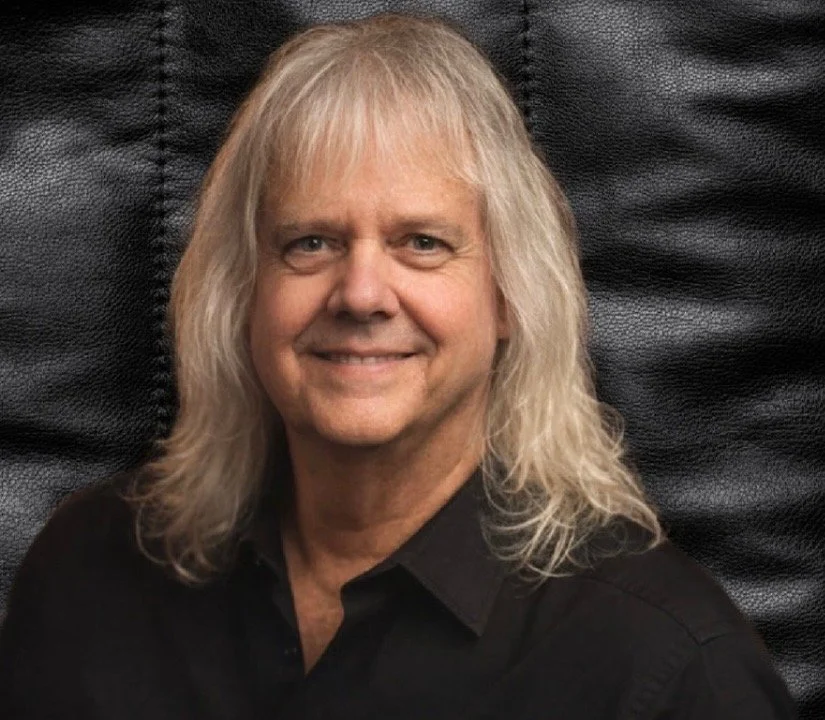 A man with long, wavy, gray hair and a friendly expression, wearing a black shirt, seated against a black textured background.