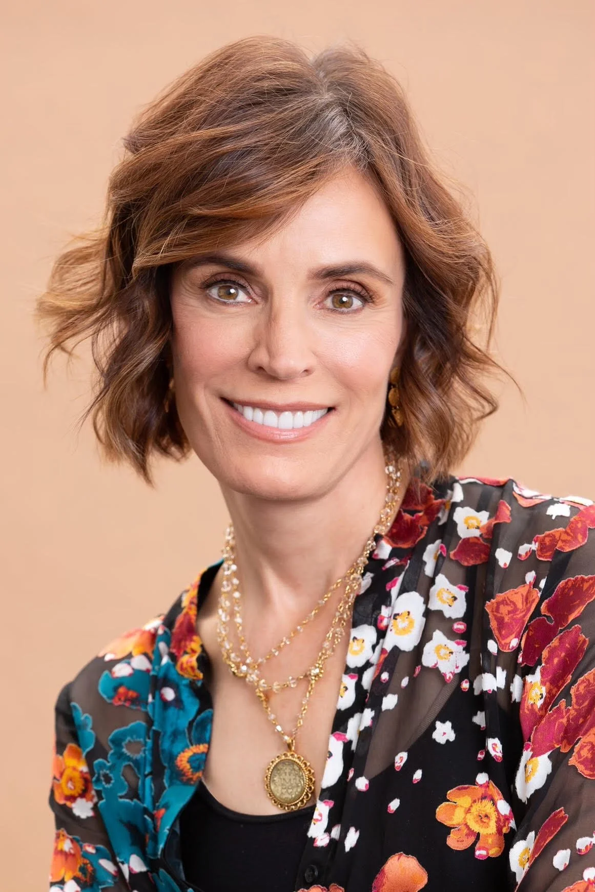 A woman with short, wavy, auburn hair and light skin, smiling, wearing a floral patterned sheer black top with various colored flowers, layered gold necklaces, and a large pendant, set against a neutral background.