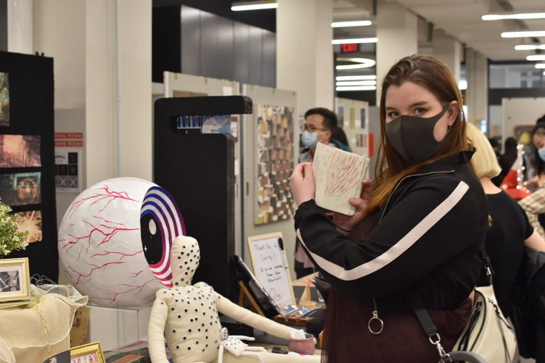 A young woman wearing a black face mask and a black jacket with white stripes on the sleeves stands at an art and craft booth in an indoor marketplace or fair. She is holding a small piece of artwork or craft in her hand, showing it toward the camera