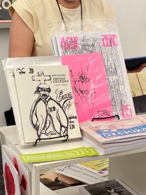 A person is holding a stack of magazines and papers with various drawings and text. On the table in front, there is a booklet with a cartoon figure wearing sunglasses and a hoodie, along with other printed materials.