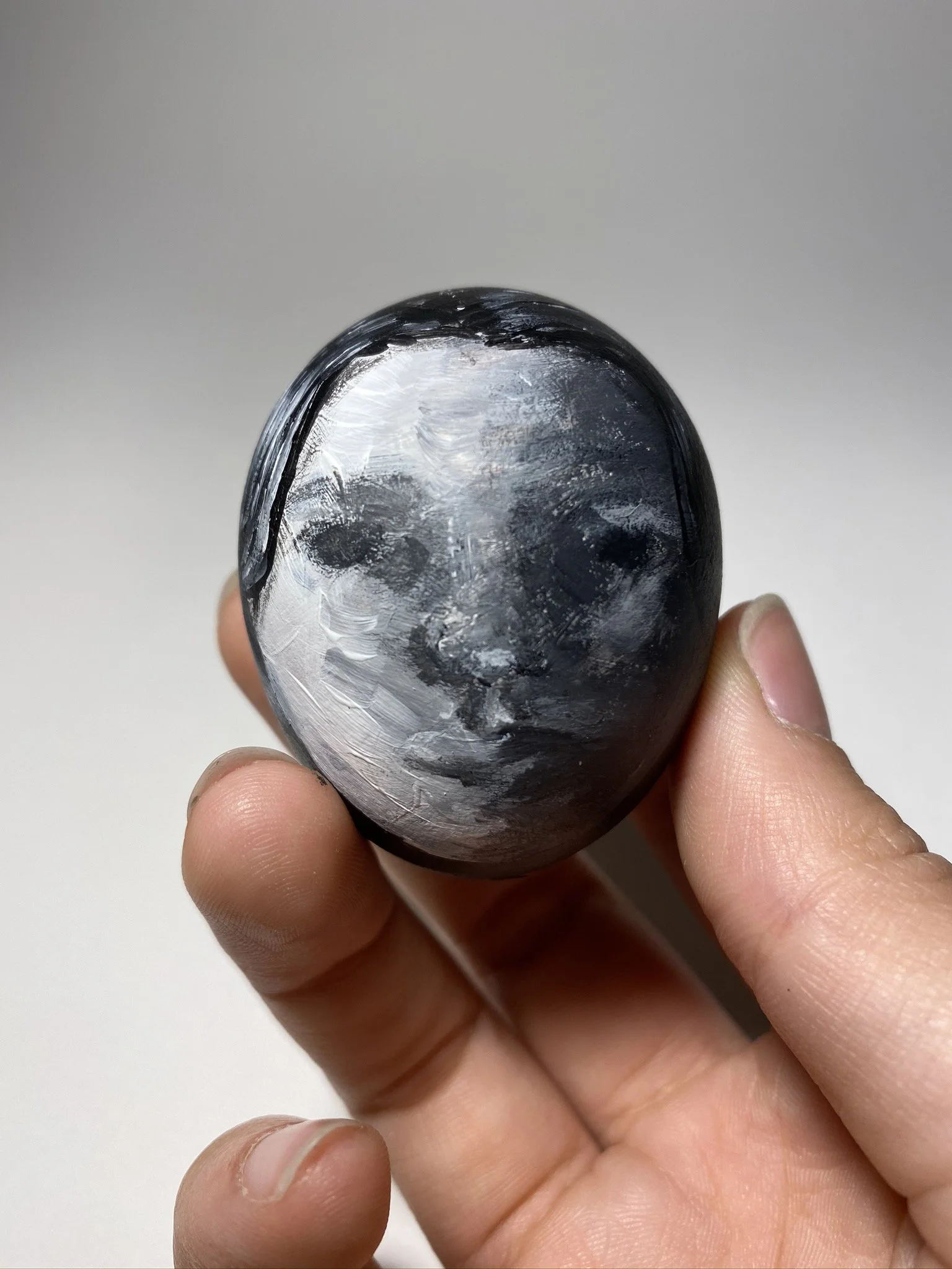 A hand holding a black and silver eggshell with a shiny, reflective surface and noticeable brush strokes, looking like a face.