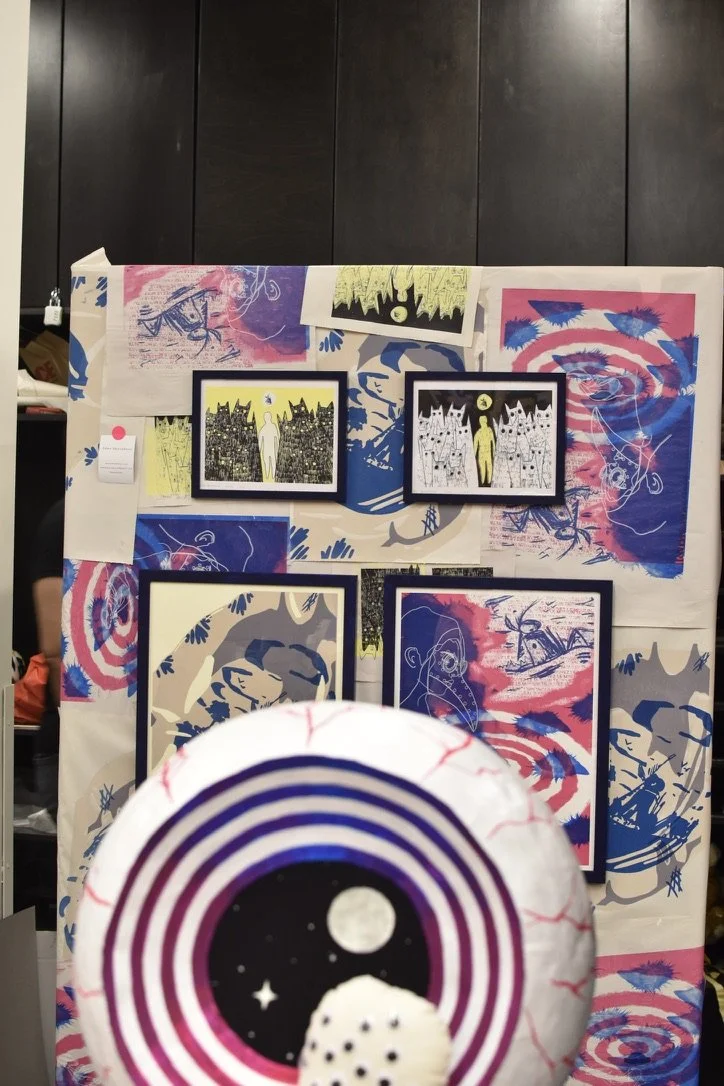 Display of various colorful abstract and black-and-white artwork and framed prints on a large white board, with a decorative lantern in the foreground featuring a cosmic eye design.