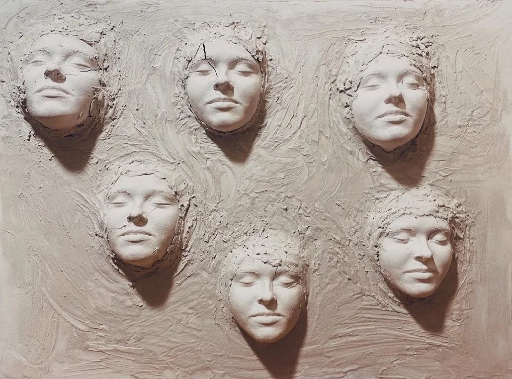 Six human face sculptures partially embedded in a textured surface, arranged in two rows of three, with serene expressions and closed eyes.