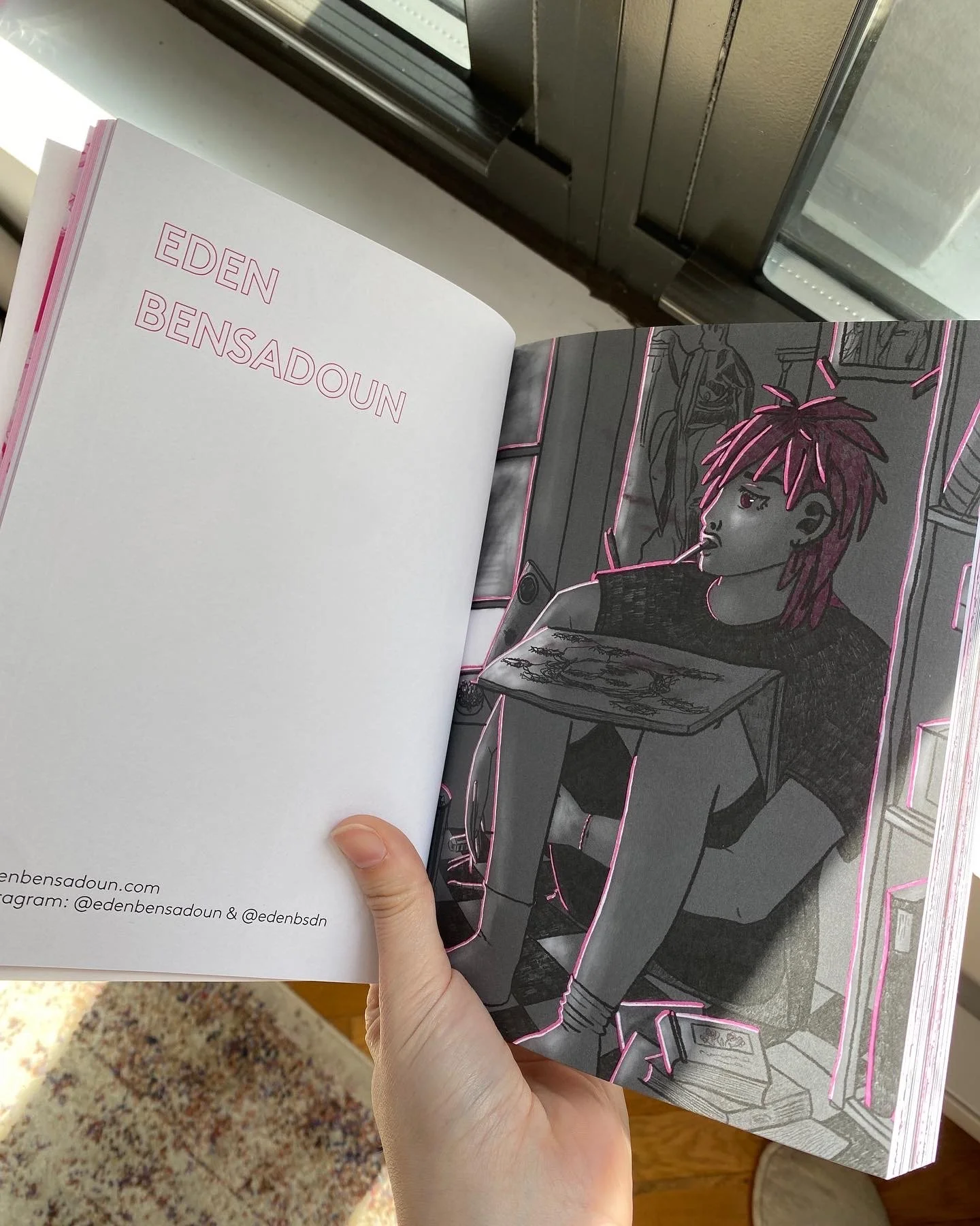 Open book displaying modern, stylized illustration of a person with pink hair, seated at a desk with scattered objetcs, in a room with framed pictures and potted plants, outlined with pink neon-like lines.