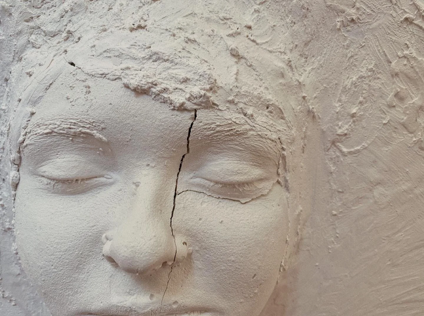 Close-up of a white plaster or ceramic sculpture of a face with closed eyes, showing cracks and textured surface.