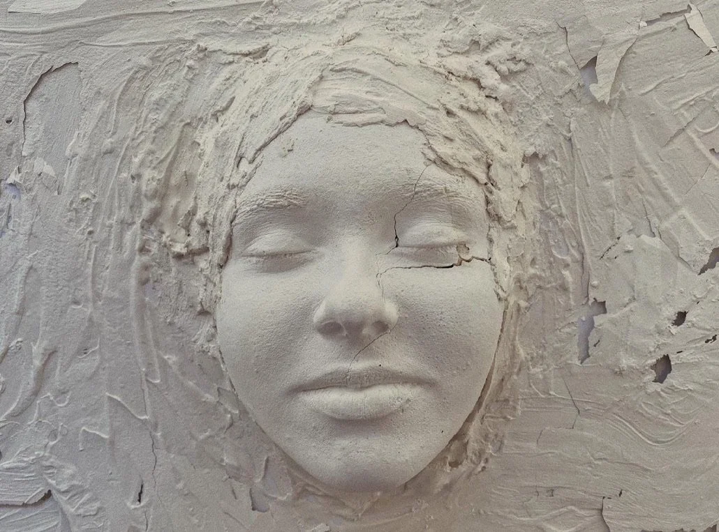 Close-up of a 3D plaster relief sculpture of a woman's face, with closed eyes and full lips, embedded in textured plaster walls.