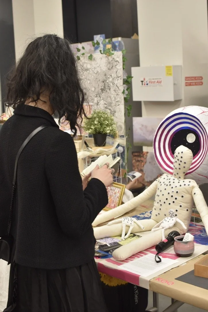 A woman with dark, curly hair wearing a black cardigan stands at a table with a plush doll with a large striped head, polka dots, and long limbs. 
