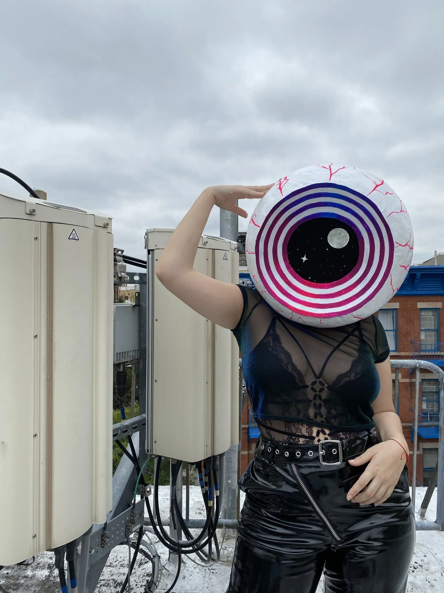 Person with a large, circular, eyeball (and cosmic-themed) headpiece and black lace top, posed next to electrical equipment on a rooftop.
