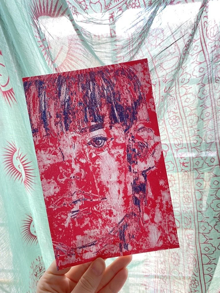 Hand holding a red and black print of a face against patterned fabric background.