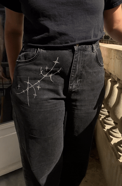 Close-up of a person wearing black jeans with a sygil constellation design bedazzled on the right thigh, and a black t-shirt, standing outdoors near stone railing.
