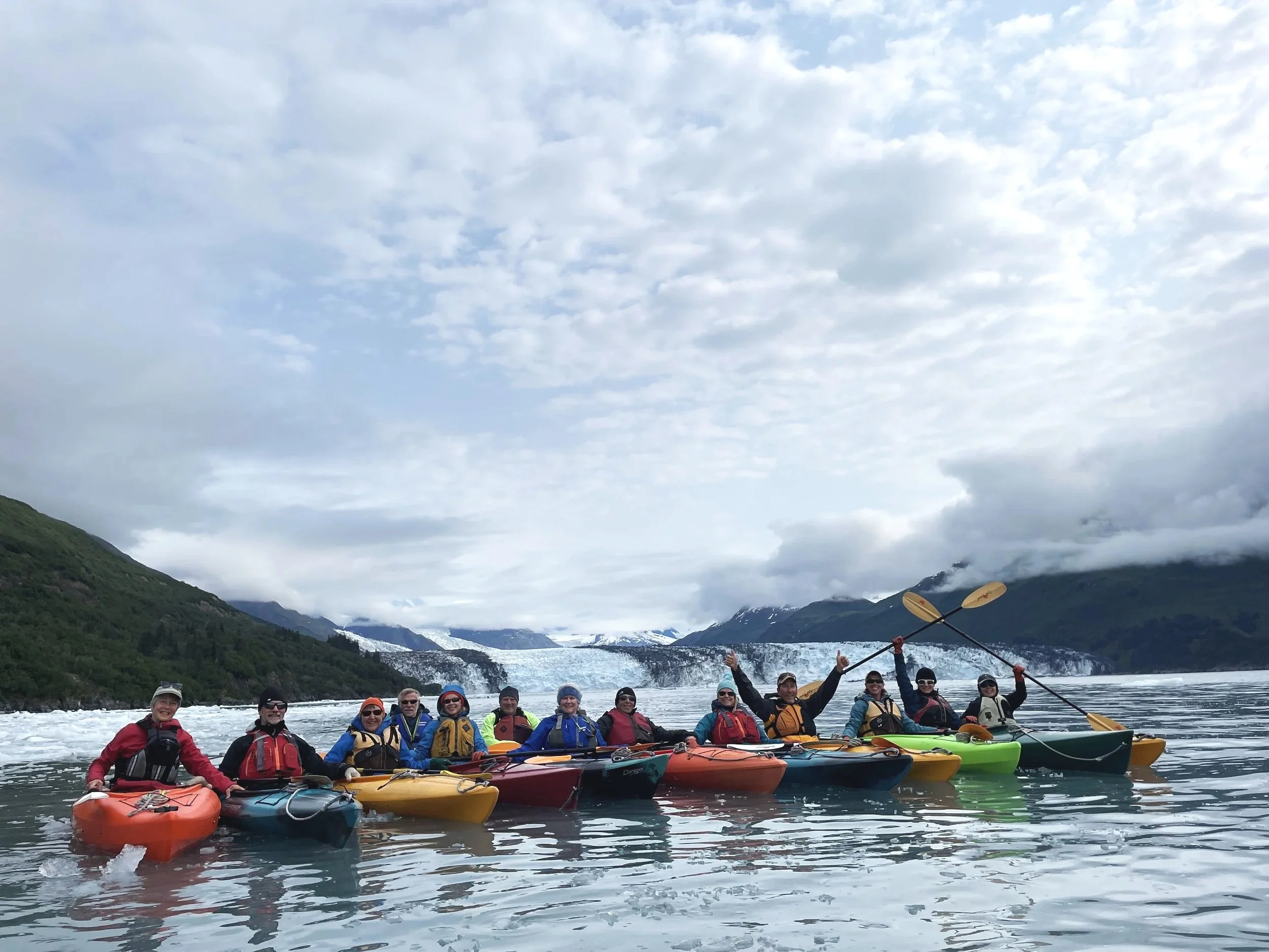 Group of adults kayaking on a river with mountains and a glacier in the background.