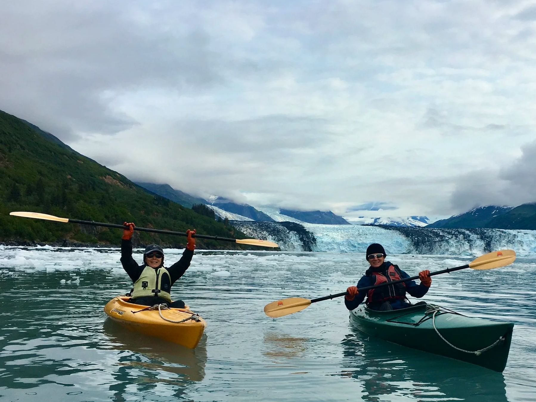 Two adults kayaking on a glacier-fed river surrounded by mountains.