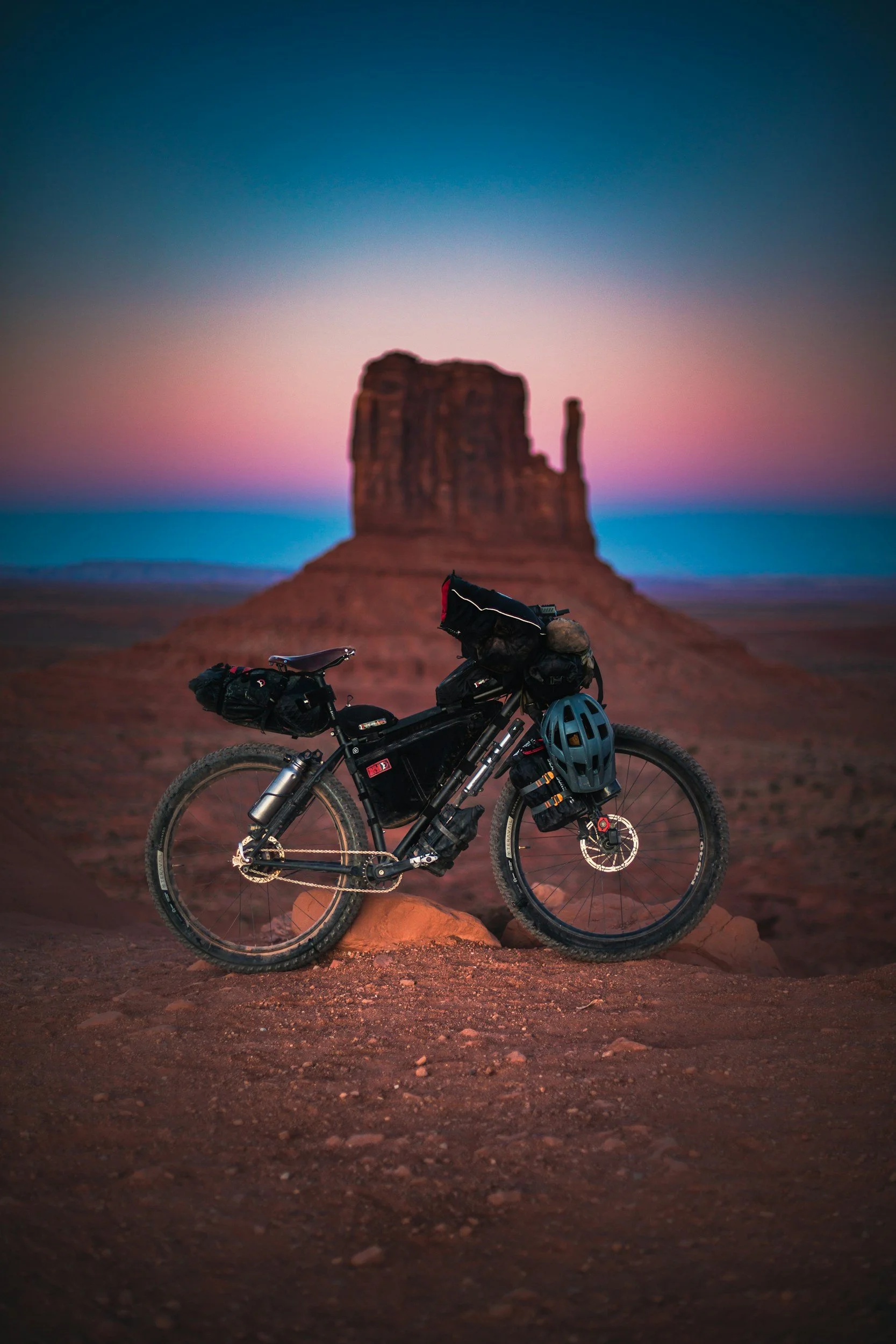 A mountain bike with gear and a helmet resting on the ground in front of the famous monument Monument Valley at sunset, with colorful sky and red rocks.