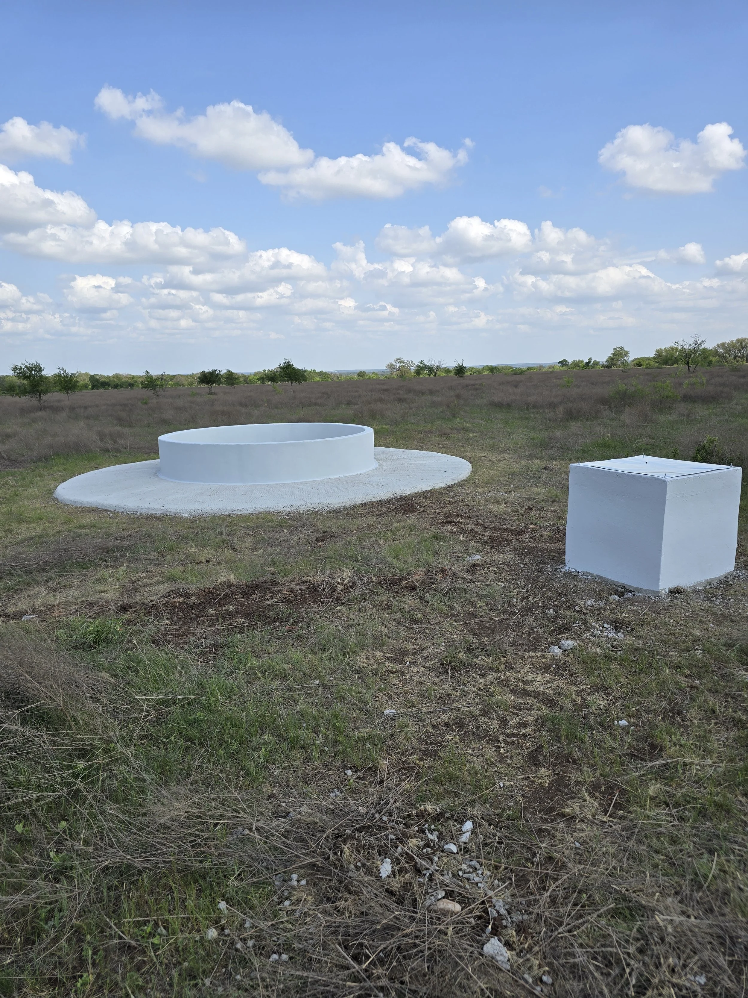 Empty white circular and square concrete structures in a grassy field with scattered trees and a blue sky with white clouds.