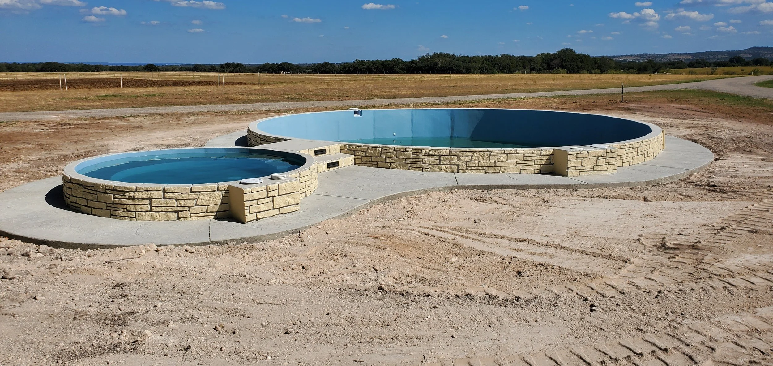 Two round hot tubs with stone exteriors, filled with water, situated outdoors on a concrete pad in a rural area with fields and trees in the background.