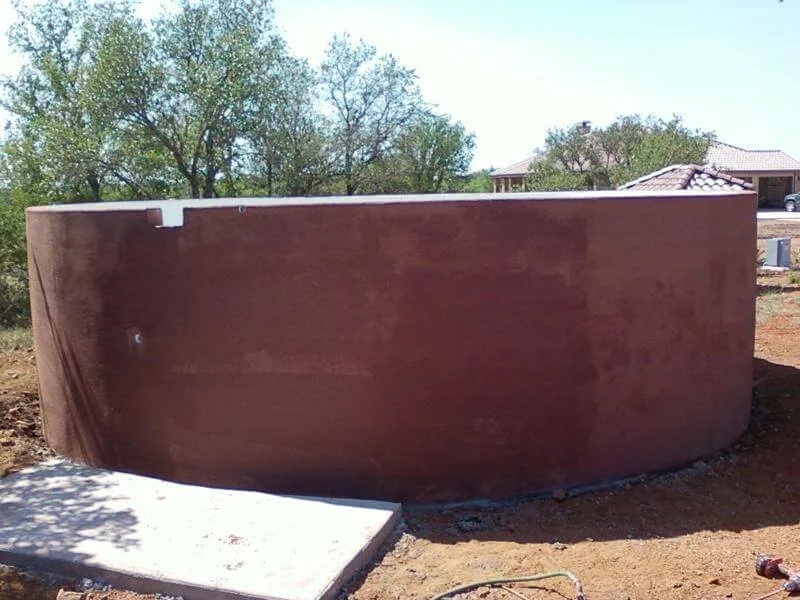 A large round, brown concrete or stucco wall under construction in an outdoor area with trees and houses in the background.