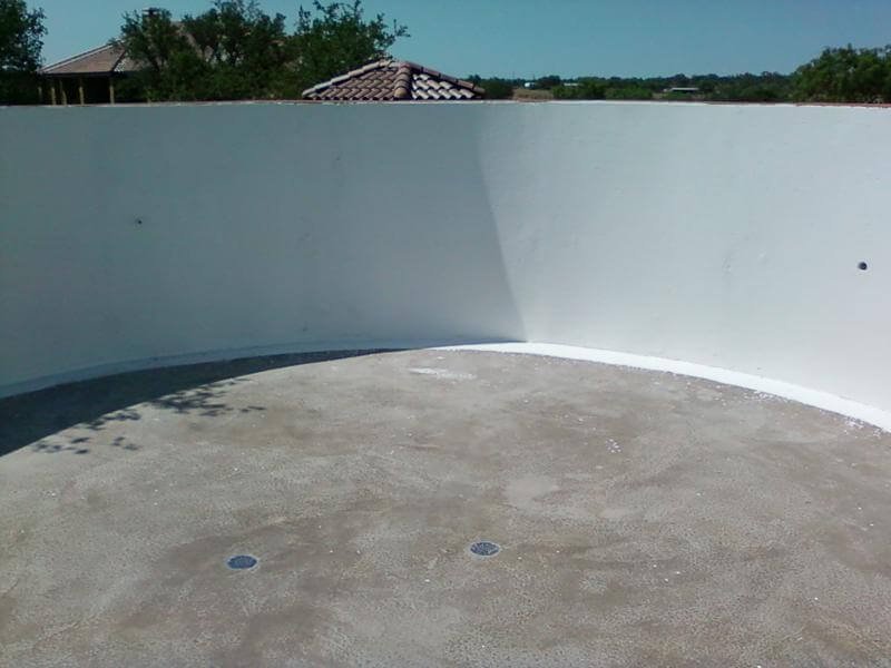 Empty rooftop terrace with a curved white wall, concrete flooring, and drainage drains, with trees and rooftops visible in the background.
