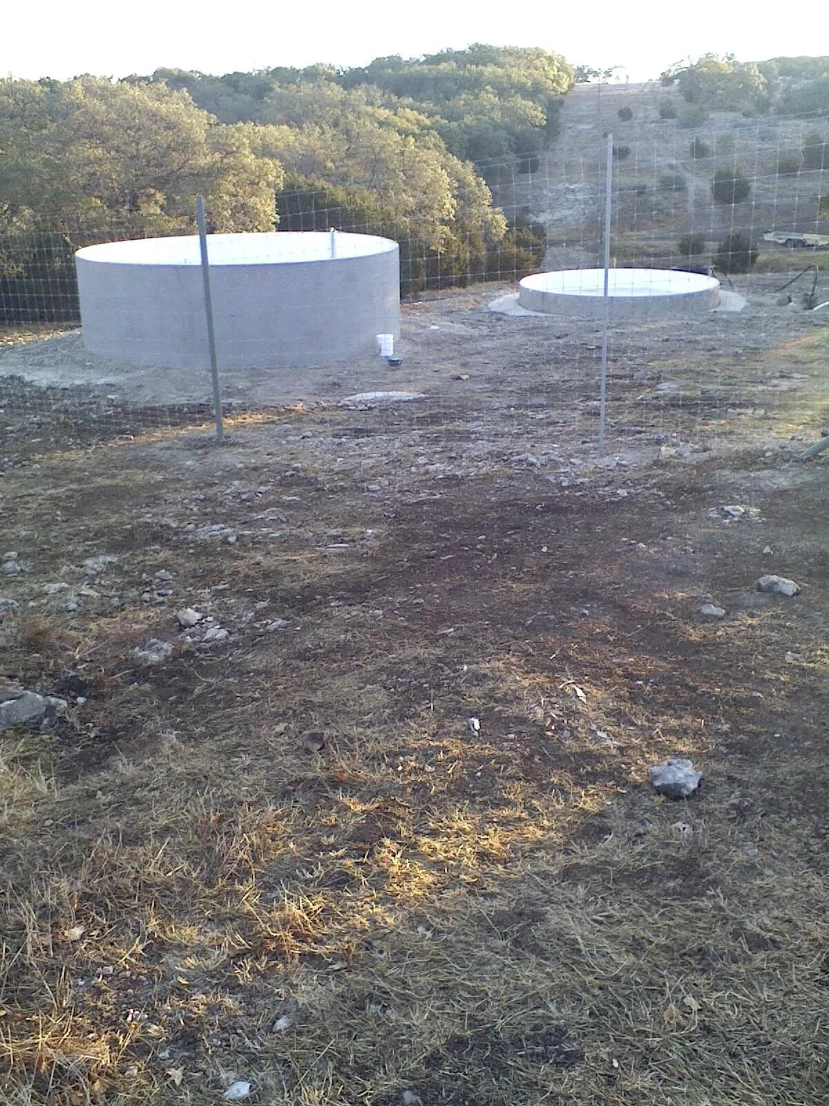 Two large round concrete structures in a dry, rocky area on a hillside, with a background of trees and a rocky cliff face, surrounded by a wire fence.