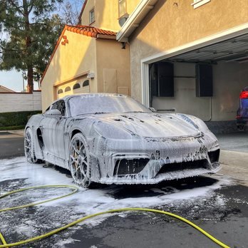 A silver sports car being washed with soap and foam on a driveway in front of a house.