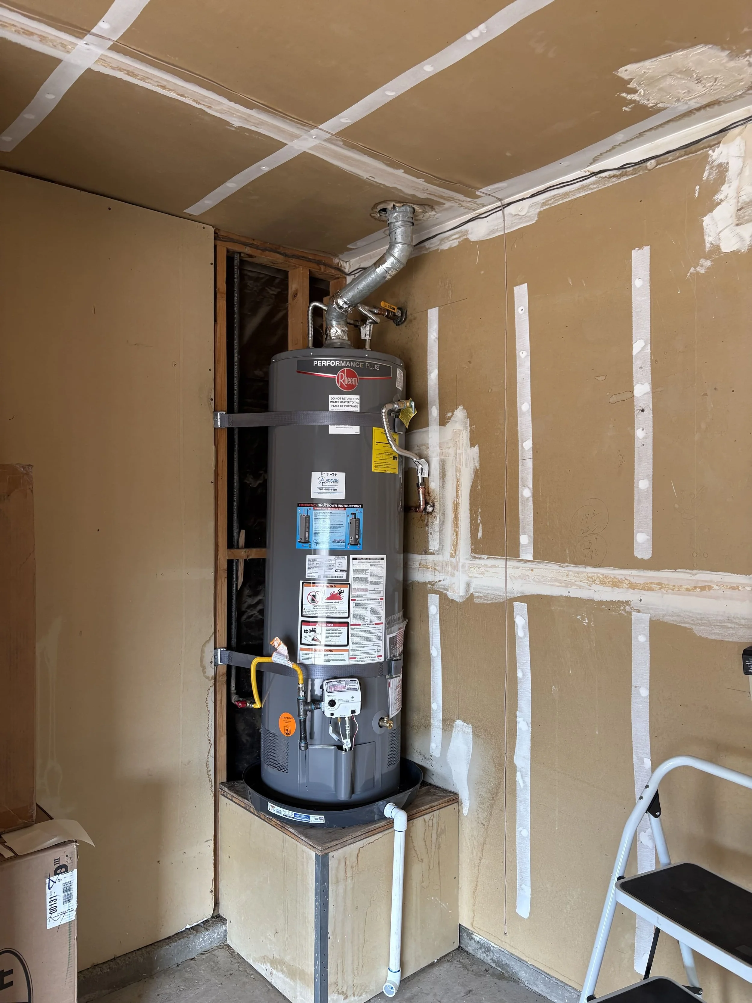 A water heater installed in a room with unfinished walls and partially visible drywall. The water heater is secured with a strap, connected to pipes, and is placed on a wooden platform with metal supports. Construction materials and tools are nearby.