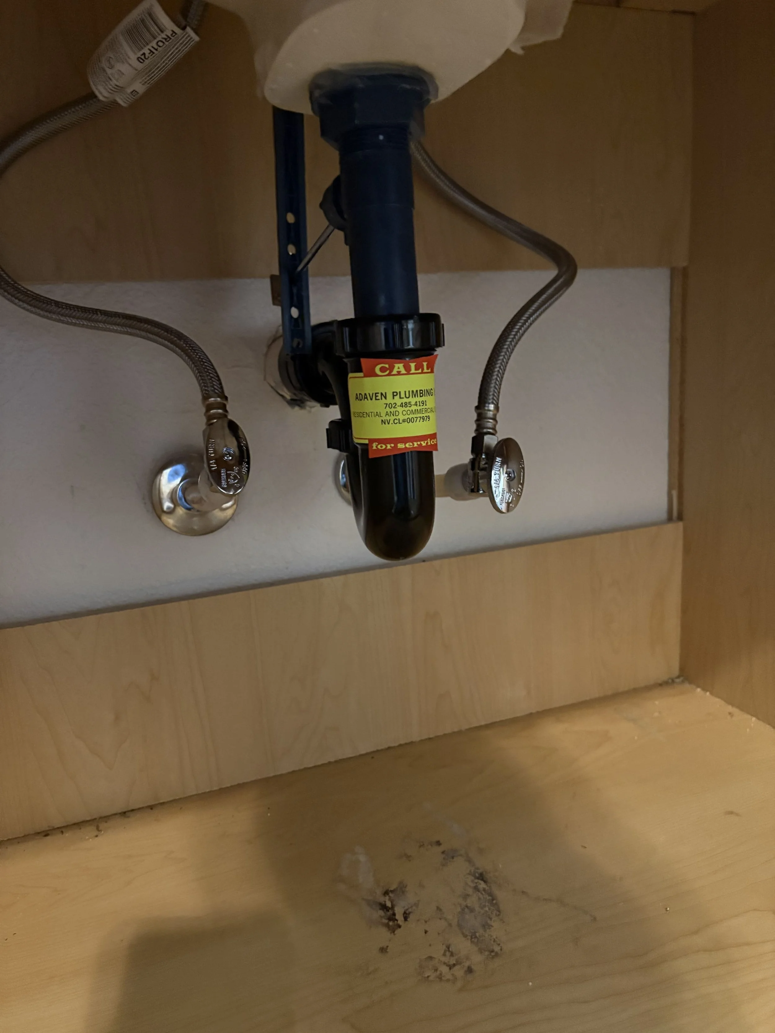 Underneath a kitchen sink, the plumbing pipe is visible with a yellow service sticker, surrounded by flexible water supply lines and some dirt on the wooden cabinet base.