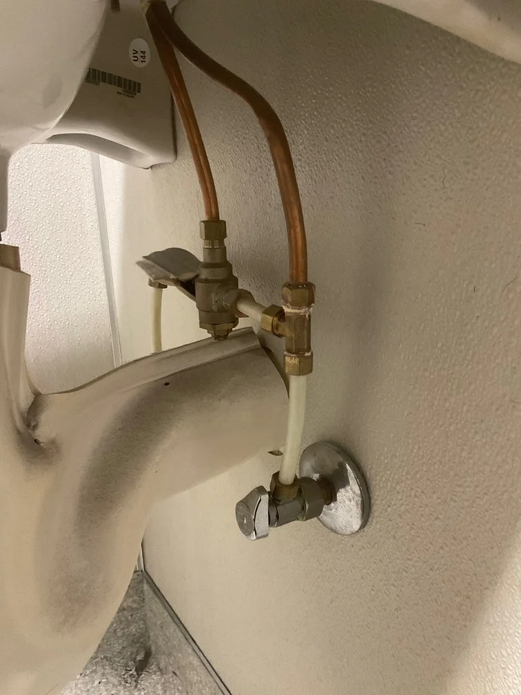 Close-up of plumbing pipes and fittings behind a urinal, including copper pipes, brass fittings, and water flowing through the pipes.