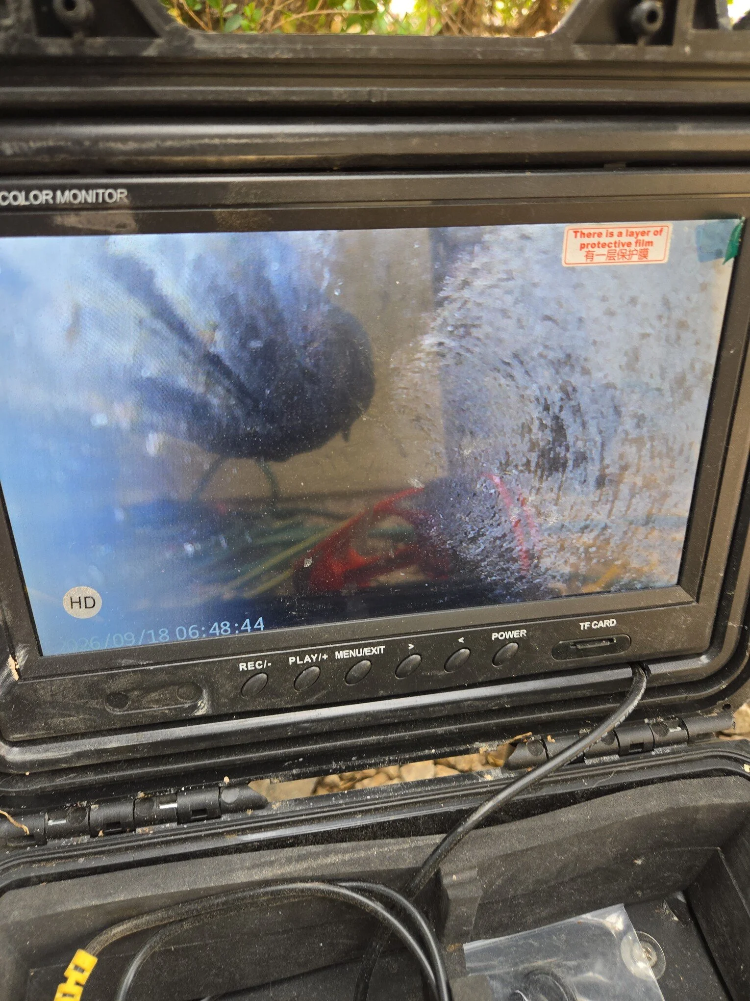 The image shows the screen of a black electronic device, possibly a monitor or a camera, with a reflective surface displaying a faint outdoor scene. There are various control buttons below the screen, including REC, PLAY, MENU/EXIT, POWER, and TF CARD. The device is situated on a black surface with visible dirt and debris, and a yellow object is partially visible in the bottom left corner. The top right corner of the screen has a red warning sticker in English and Chinese stating, 'There is a layer of protective film'.