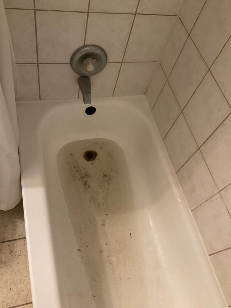 A dirty bathtub with rust and grime, with a wall-mounted showerhead above and tiled walls around it.