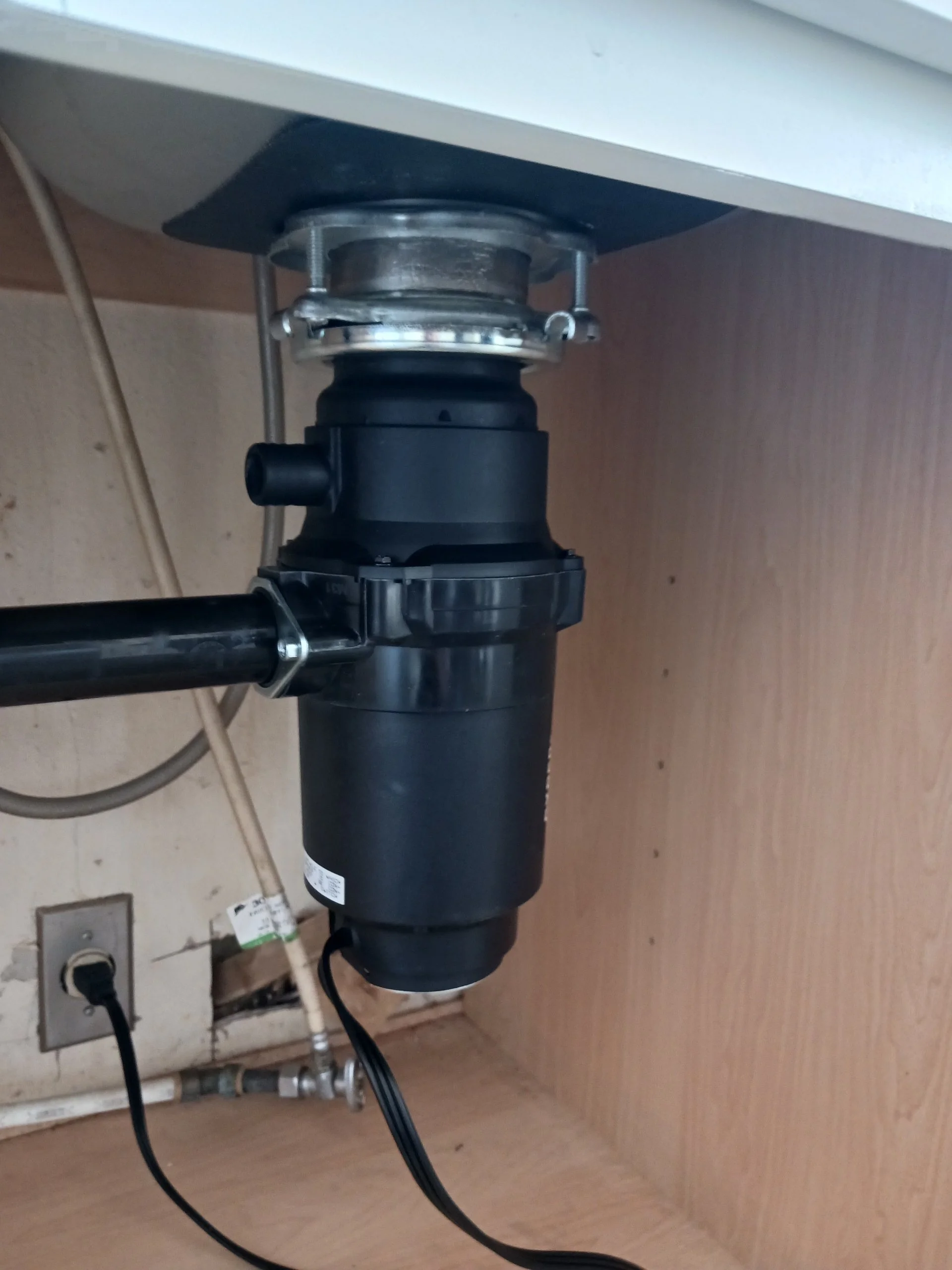 Under-sink garbage disposal unit connected to plumbing and electrical outlet in a kitchen cabinet.