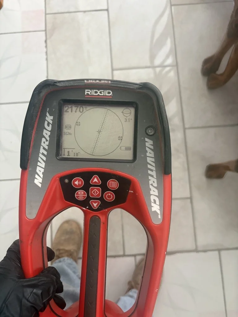 A person wearing a black glove holding a RED RIDGID Navitrack metal detector, which displays a circular readout showing a depth of 11 inches and a target signal strength of 2170. The person is standing indoors on a tiled floor, with two dog paws visible on the right side.