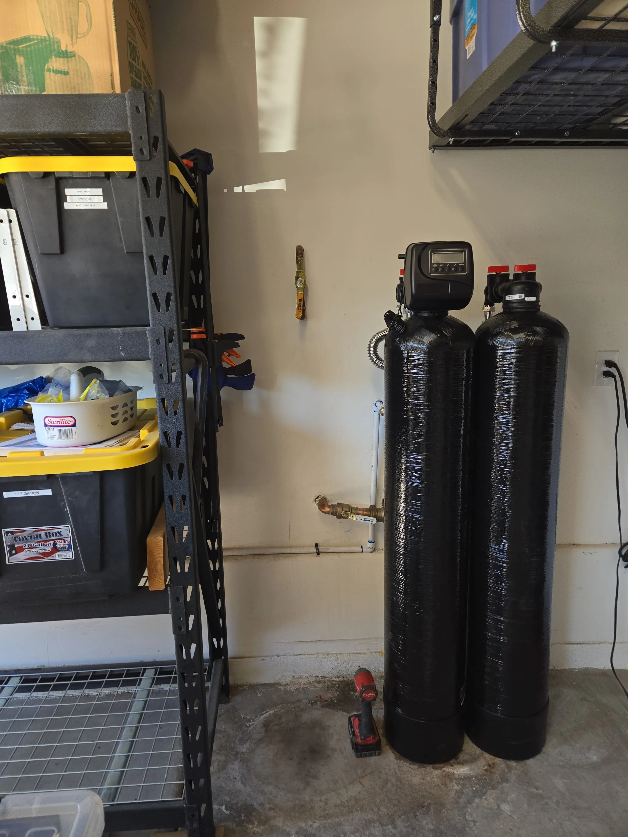 Garage utility room with two large black water softener tanks, a digital control panel on top, and connected pipes. Shelving with storage bins and tools, ladder, and various small items are also visible.