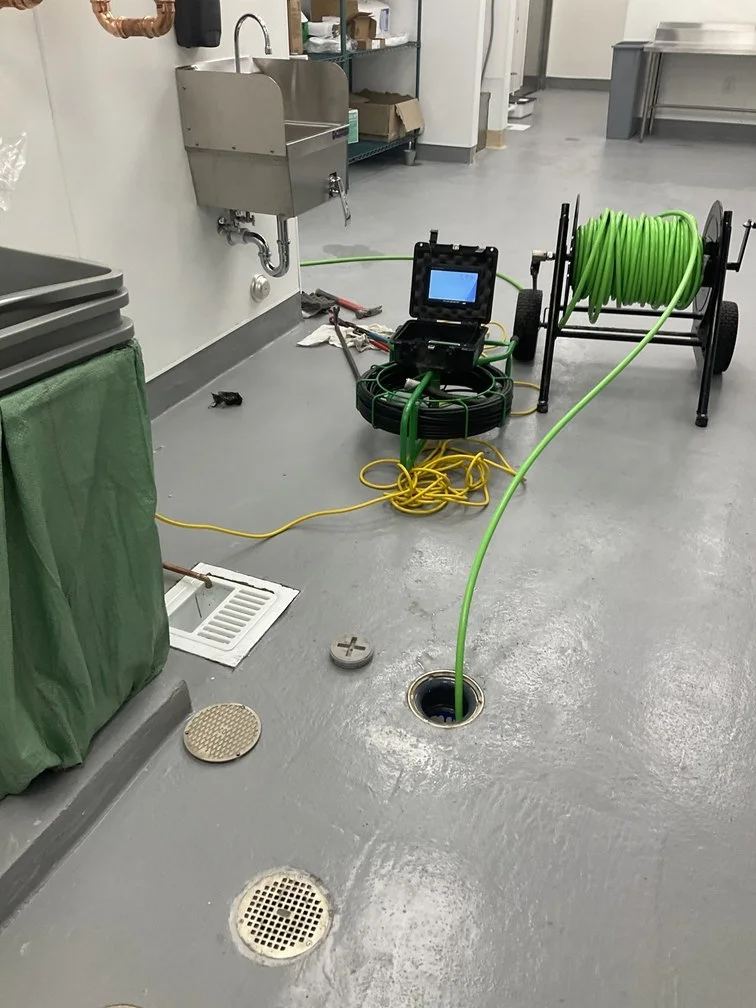 Industrial cleaning equipment with green hoses on a reel, a small monitor, and drainage fixtures in a commercial or laboratory setting.