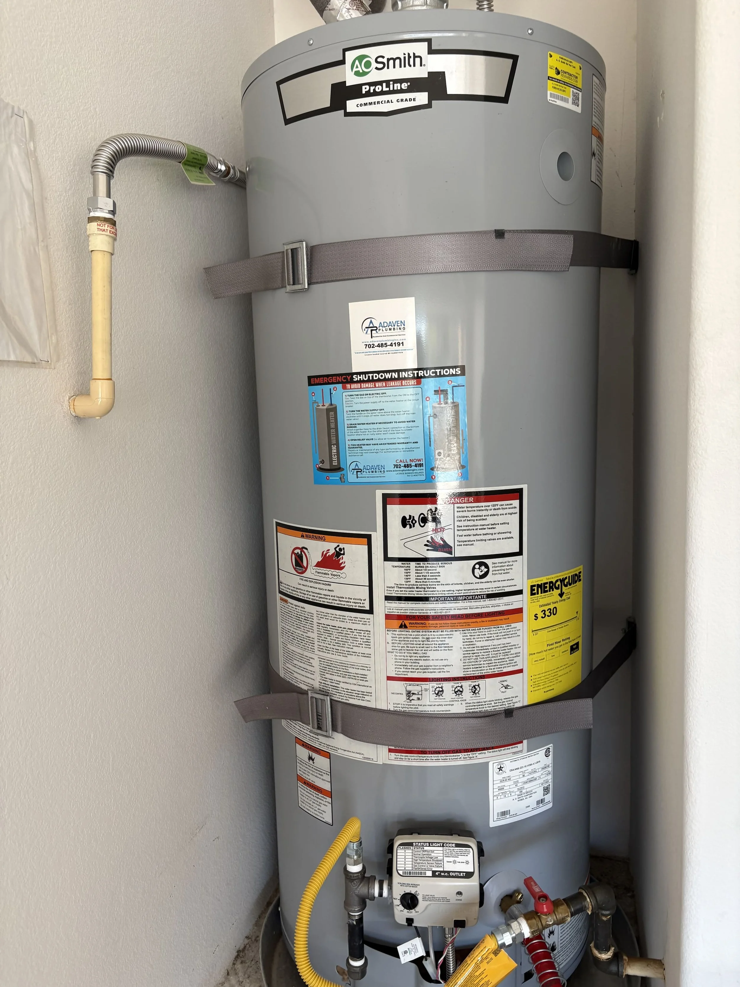 A large grey water heater with various labels and warning signs, connected to pipes and fittings, installed in a corner of a room.